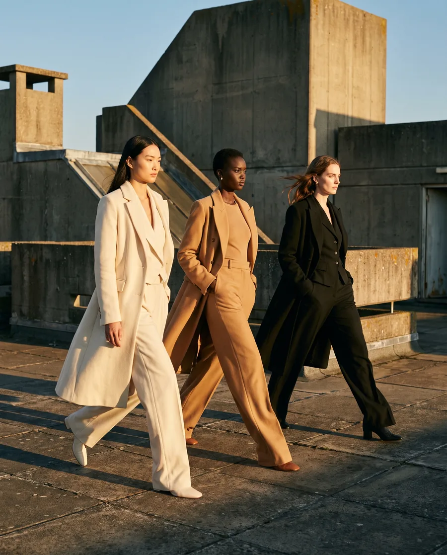 Three models in cream, camel and black walk across a concrete rooftop in low sunlight